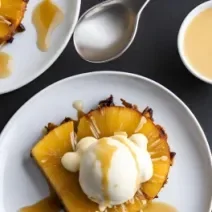 Fotografia vista de cima de fatias de abacaxi caramelizado com uma bola de sorvete de creme e chocolate branco derretido, sobre um prato branco. Ao lado tem outro prato com a mesma sobremesa e pote pequeno branco com galak derretido.