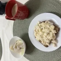 Fotografia de um prato raso médio branco com uma fatia de pão e ovos mexidos cremosos com farelo de aveia. Ao lado tem um recipiente pequeno de vidro e branco com melão e aveia e uma xícara vermelha grande.