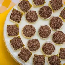 Fotografia em tons de amarelo em uma bancada de madeira com um pano amarelo e outro amarelo xadrez com branco. Em cima deles, um suporte para bolo branco com vários brigadeiros em forminhas amarelas.