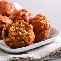 Fotografia de alguns muffins de maçã sobre um apoio retangular branco, o qual está apoiado em um paninho também branco. O pano está sobre uma bancada de cimento queimado claro.