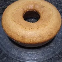 Fotografia de um bolo de fubá, feito na forma de furo central, apoiado em um vidro redondo, sobre uma bancada de cor escura.