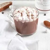 Fotografia em tons de marrom em uma mesa de madeira branca, um pano branco e uma xícara de vidro com o chocolate quente dentro e minis marshmallow para decorar a xícara. Ao lado, uma colherzinha de madeira.