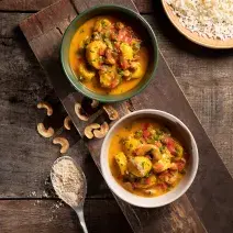 Foto de cima de dois pratos redondos e fundos com a receita de moqueca de banana da terra, ambos sobre uma tábua de madeira, com algumas castanhas-de-caju na mesa, uma colher de metal com arroz e um prato com arroz branco