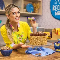 Foto da confeiteira e influenciadora Beca Milano atrás de uma bancada de cozinha com o Bolo Charge e alguns bombons Charge