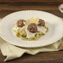 Fotografia em tons de marrom e branco de uma bancada marrom vista de cima. Contém um pano pardo com um prato branco ao centro com macarrão e bolinhos de carne.