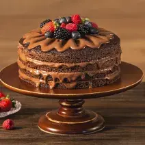 Fotografia em tons de marrom em uma bancada de madeira escura com um suporte para bolo marrom e o naked cake com ganache de cacau e frutas vermelhas em cima. Ao lado, um pratinho de vidro com frutas vermelhas.