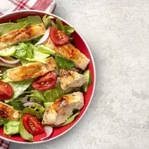 Fotografia vista de cima de um salada de folhas pepino, tomate, frango, cebola roxa e salsinha com um molho de maracujá e mel. A salada está dentro de um recipiente fundo vermelho, apoiado sobre e metade de um pano branco com linhas vermelhas.