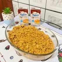 Foto da receita de ambrosia em tom amarelo, numa travessa de vidro, sobre uma mesa com toalha e nela há duas caixas do produto de Moça e uma vasinho com uma planta em tom verde