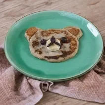 Foto da receita de Panqueca de Banana. Observa-se um pratinho verde e a panquequinha com cara de ursinho.