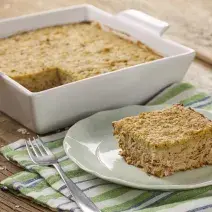 Fotografia em tons de branco e dourado, refratário branco com torta de frango, prato branco com uma porção de torta de frango com um garfo ao lado e sobre guardanapo com listras verdes, azuis e brancas, colher e potinho com aveia ao fundo.