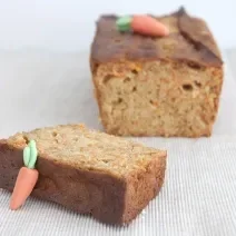 Fotografia em tons claros de um bolo de cenoura com mini cenouras em cima, feitas de pasta americana, cortado em uma fatia grossa que está mais no centro da foto. O bolo está sobre uma toalha de mesa na cor marrom claro.