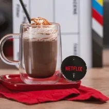 Foto em tons de vermelho e preto da receita de cappuccino netflix servida em um copo largo sobre uma base de porcelana vermelha e um pano vermelho em cima de uma mesa de madeira com uma cápsula de cappuccino netflix ao lado