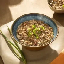 Foto em tons de marrom da receita de risoto de filé mignon e gorgonzola servida em um bowl de cerâmica azul sore um pano bege com ramos de alho poró ao lado