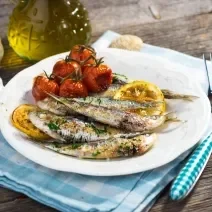 Fotografia em tons de branco com um prato branco e redondo ao centro. Em cima do prato existe sardinhas assadas com tomates e limão siciliano. Ao lado existe um garfo e uma faca de cor prateada.