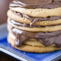 Fotografia de biscoitos empilhados, e entre alguns, um creme de chocolate caindo um pouco nas bordas. Os biscoitos estão em um prato quadrado na cor azul em tom mais escuro, que estão sobre uma mesa na cor marrom escuro.