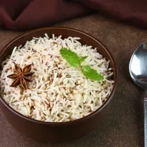 Fotografia em tons escuros de um recipiente fundo na cor marrom com arroz e especiarias e duas folhas de hortelã por cima. Ao lado, uma colher de sopa desenhada, sobre uma mesa marrom em tom escuro.