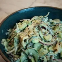 Fotografia em tons de marrom escuro com um bowl escuro ao centro. Dentro do bowl existe uma receita de salada de pepino feita com cream cheese, salmão, cebola e gergelim preto