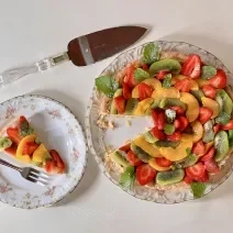 Foto vista de cima da receita de torta de frutas com creme de amêndoa, servida em um prato redondo ao lado de uma fatia cortada. A torta é decorada com frutas diversas (kiwi, manga e morango)