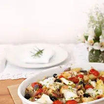 Fotografia em tons de branco e vermelho de uma bancada de madeira vista de lado, ao centro um recipiente oval branco, dentro dele pedaços de bacalhau, tomates cereja e azeitonas. Ao fundo um prato branco e um guardanapo para decorar.