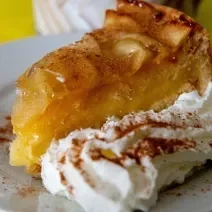 Fotografia em tons de branco e amarelo com um prato branco ao centro. Dentro do prato existe um bolo de maçã polvilhado com canela acompanhado de um jato de chantilly.