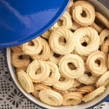 Fotografia em tom claro, com biscoitos amanteigados em formato redondo, modelado com bico de confeitar trançado, dentro de um refratário. No canto superior esquerdo da foto, uma tampa azul.