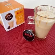 Fotografia em tons de laranja e vermelho com um copo de vidro ao centro. Dentro do copo existe uma receita de cappuccino com calda de caramelo e ao lado uma caixa de cápsulas Dolce Gusto Cappuccino NETFLIX Caramelo Salgado