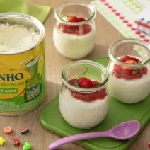 Fotografia de três recipientes de vidro pequenos com pudim de tapioca de ninho um creme de morango por cima. Dois desses recipientes estão sobre um apoio verde retangular, ao lado de uma colher roxa pequena e de uma lata de ninho escolar.