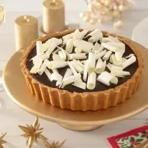 Foto da receita de Torta de Biscoito com Ganache de Café. Observa-se uma torta inteira sobre uma boleira dourada decorada com raspas de chocolate branco por cima e decorações de festa atrás.