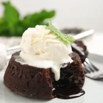 Fotografia de um petit gateau com uma bola de sorvete por cima e uma folha de hortelã. A sobremesa está em um prato raso e grande, com um garfo segurando um pedaço do bolinho e uma colher de sorvete meio cheia.