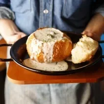Fotografia de um chef segurando uma tábua de madeira, com uma panela grande preta em cima, com um pão aberto recheado com um creme de palmito que está transbordando e caindo na panela. Por cima, salsinha picada e salpicada.