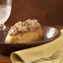 Fotografia em tons de marrom e pardo, em uma bancada contém um pano cor pardo com um garfo por cima, um recipiente marrom com uma batata com pedaços de carne com molho por cima e ao lado um copo transparente com agua