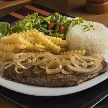 Fotografia em tons de marrom, branco e amarelo de uma bancada marrom, contém um prato redondo e branco com um bife com cebolas por cima, arroz, batatas fritas e salada como acompanhamento