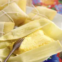 Fotografia em tons de amarelo em uma bancada de madeira, uma toalha colorida florida e algumas pamonhas de queijo coalho e alecrim em cima da bancada.