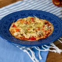 Fotografia em tons de azul com um prato azul ao centro. Em cima do prato existe uma receita de pizza de frigideira feita com frango desfiado, molho de tomate, queijo muçarela e orégano.