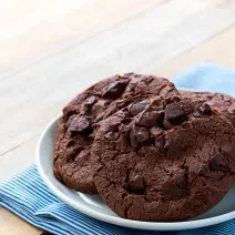 Fotografia de quatro cookies de chocolate com gotas de chocolate dentro de um recipiente de vidro branco um pouco fundo. O recipiente está por cima de um paninho azul com listras brancas sobre uma mesa de madeira.