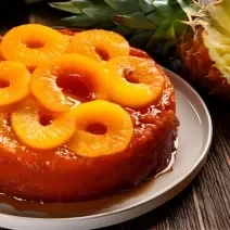 Fotografia de um bolo de abacaxi inteiro assado em forma redonda sem furo. Por cima do bolo tem caramelo e rodelas de abacaxi, e o bolo está apoiado em um prato raso grande de tom claro sobre uma mesa de madeira, ao lado de uma metade de abacaxi.