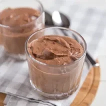 Receita de Mousse de Chocolate da Tia Alzira, tradicional e prático, feito com Chocolate Meio Amargo NESTLÉ CLASSIC, NESTLÉ Creme de Leite e claras em neve, servido em copinhos sobre uma mesa com uma tábua de madeira e colheres ao fundo