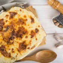 Fotografia de uma travessa de vidro quadrada de bacalhau gratinado com batata sobre uma tábua de madeira e um pano de tom cinza. No fundo tem um moedor de pimenta de madeira.