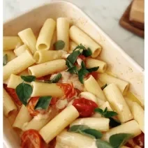 Fotografia de uma assadeira de cor clara com macarrão penne, tomates cereja assados, queijo feta derretido e folhas de manjericão.