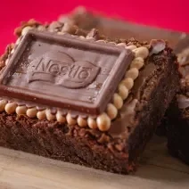 Fotografia três unidades de brownies com biscoito choco biscuit, eles estão apoiados um no outro e sobre uma tábua de madeira. O fundo da foto é vermelho.