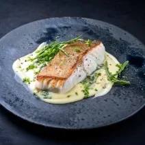 Fotografia um filé de pescada assado sobre um creme de legumes em um prato raso escuro, o qual está sobre uma mesa preta.