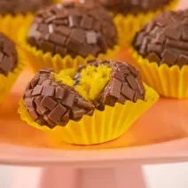 Fotografia em tons de amarelo, rosa e marrom. Ao centro um brigadeiro mordido, mostrando o recheio de bolo de cenoura.