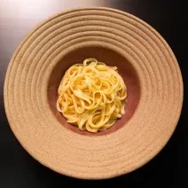 Fotografia em tons de cinza vermelho com um prato vermelho claro ao centro. Dentro do prato existe uma receita de fettuccine Alfredo feita com manteiga e queijo.