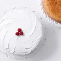 Fotografia em tons de branco e dourado, com bolo decorado com glacê branco e decorado com três framboesas sobre prato de vidro, ao lado massa de pão de ló assada sobre prato de vidro, tudo sobre bancada em tom de cinza.