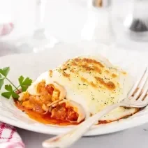 Fotografia em tons de branco de um canelone recheado de abóbora com carne seca, e por cima, um molho de requeijão gratinado no forno.