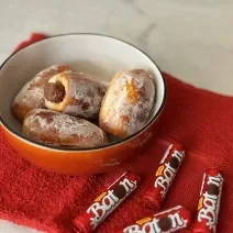 Fotografia mostra um pão tipo bisnaguinha recheado com chocolate baton. Os pãoszinhos estão dentro de um bowl vermelho. Ao lado, batons ao leite dispostos sobre um pano vermelho.