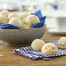 Foto de uma mesa com um pano e 3 pães de queijo redondos em cima. No fundo da foto há uma tigela com mais pães dentro