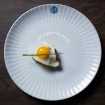 Fotografia vista de cima de um prato branco com steak tartare, chips de batata e ovo de codorna frito por cima, decorado com cebolete. O prato está sobre uma bancada de madeira.