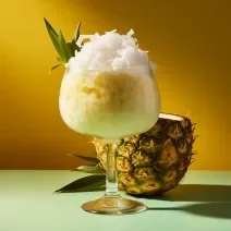 Fotografia de uma taça grande de vidro com gelado de abacaxi e coco, com lascas de coco por cima. A taça está perto de metade de um abacaxi cortado.