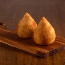 Fotografia de duas coxinhas de frango sobre uma tábua pequena de madeira. A tábua está sobre uma mesa de madeira.